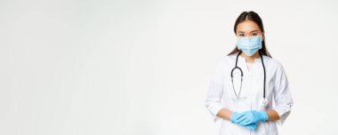 Covid-19 ve sağlık hizmeti konsepti. Tıbbi maskeli genç Asyalı kadın doktor, lastik eldivenler ve klinik üniforması yardım etmeye hazır, dinleyen hasta, beyaz bir geçmişi var..