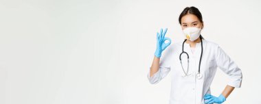 Gülümseyen Asyalı doktor, kadın doktor iyi olduğunu gösteriyor, solunum cihazı takıyor, solunum cihazı takıyor, beyaz arka planda üniforma içinde duruyor..