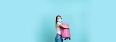 Mutlu Asyalı kadın turist, bavuluyla tıbbi maske takarak dans ediyor, tatile çıkıyor, evrak çantasıyla seyahat ediyor, mavi arka planda duruyor..