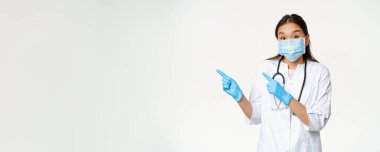 Heyecanlı Asyalı kadın doktor solu işaret ediyor, önleyici tedbir olarak tıbbi maske takıyor, beyaz arka planda üniforma giyiyor..