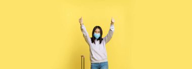 Asyalı mutlu bir turist, tıbbi maske takan bir kız, başparmağını kaldırıyor ve bavulun yanında duruyor, sarı arka plan..