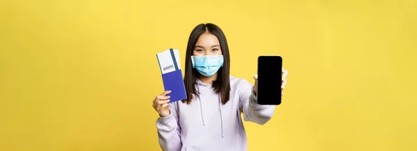 Happy korean girl in face mask, showing smartphone screen, app interface and passport with tickets for vacation, booking trip, yellow background.