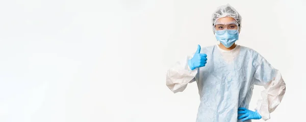 Asian female doctor or nurse wearing personal protective equipment and rubber gloves, medical face mask, showing thumbs up, vaccination and covid-19 campaign concept.