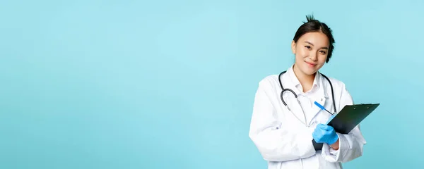Portrait of female doctor, asian physician in medical uniform, writing down patient info, standing against blue background. Healthcare concept