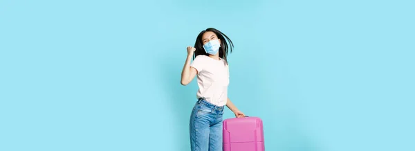 Full length of happy asia female tourist, dancing and jumping with suitcase in medical face mask, excited about vacation, standing over blue background.