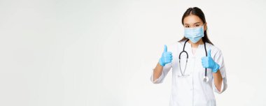 Covid-19 ve sağlık çalışanları konsepti. Yüzünde maske ve lastik eldiven olan gülümseyen Asyalı kadın doktor baş parmak gösteriyor, beyaz arkaplan.