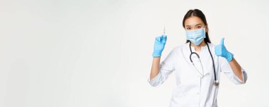 Covid-19 aşısı ve sağlık sigortası. Tıbbi maske ve eldiven takan Asyalı kadın doktor elinde aşı şırıngası ve baş parmağı havada, beyaz arka plan.