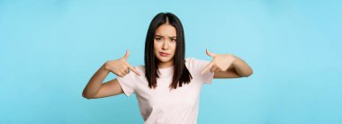 Şımarık ve havalı Asyalı kadın logoyu işaret ediyor, pürüzsüz ve çizik kaşları gösteriyor, mavi arka planda duruyor..