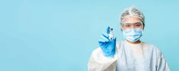 Covid-19 vaccination concept. Asian female nurse or doctor in personal protective equipment, showing coronavirus vaccine bottle, standing over blue background.
