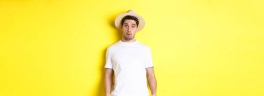 Hasır şapkalı kafası karışmış gezgin kafası karışmış, kameraya bakıyor, sarı arka planda dikiliyor. Boşluğu kopyala