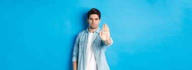 Ciddi bir adam kaşlarını çatmış ve hayır diyor, el pençe divan, dur işareti, eylemi yasaklıyor, mavi arka planda duruyor..