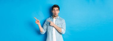 Gayri resmi kıyafetli, şaşırmış sakallı adamın portresi. Parmakla göstererek harika bir tanıtım teklifi sunuyor. Mavi arka planda duruyor..