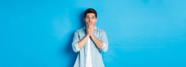Şok olmuş yetişkin bir adamın görüntüsü kameraya hayranlıkla bakıyor, nefes nefese ve ağızlarını ellerle kapatıyor, mavi arka planda dikiliyor..
