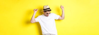 Turizm, seyahat ve tatil konsepti. Erkek turist tatilde, hasır şapka ve güneş gözlükleriyle dans ediyor, sarı arka planda poz veriyor..