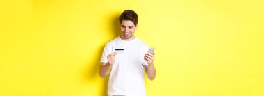 İnternetten sipariş veren adam, cep telefonu başvurusunda kredi kartı kaydı, akıllı telefonu tutan ve gülümseyen, sarı arka planda duran..