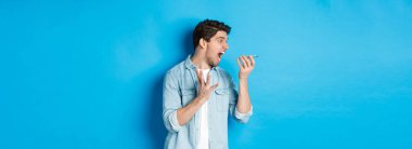 Sinirli ve sinirli bir adam cep telefonuna bağırıyor, hoparlörden bağırıyor ve mavi arka planda duruyor..