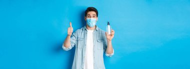 Covid-19 kavramı, salgın hastalık ve sosyalleşme. Tıbbi maskeli neşeli bir adam, el dezenfektanı ve baş parmağını gösteriyor, dezenfektan öneriyor, mavi arka plana karşı duruyor..