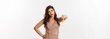 Şık elbiseli, kuşkucu genç kadın hayal kırıklığına uğramış görünüyor, baş parmağını kaldırıyor, Noel partisinden hoşlanmıyor, beyaz arka planda duruyor..