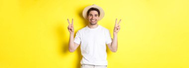 Turizm ve tatil kavramı. Barış işaretleriyle poz veren mutlu erkek turist, heyecanla gülümsüyor, sarı arka planda duruyor..