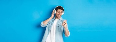 Kulaklıkla müzik dinleyen ve akıllı telefondan mesaj okuyan mutlu bir adam. Heyecanlı bir şekilde gülümsüyor, mavi arka planda duruyor..