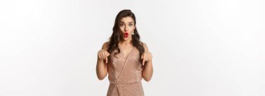 Parti elbisesi içinde şaşaalı bir kız, şaşkın bir şekilde, yılbaşı reklamını gösteriyor, beyaz arka planda duruyor..