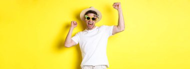 Turizm ve yaşam tarzı kavramı. Mutlu şanslı adam seyahati kazanıyor, bayram kıyafeti giyiyor, şapkası ve güneş gözlüğü takıyor, sarı arka plan.