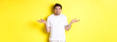 Hiçbir şeyden haberi olmayan adam omuz silkiyor, ellerini yana açıyor. Hiçbir şey bilmiyor, sarı arka planda duruyor..