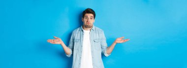 Kafası karışmış genç adam omuzlarını silkiyor, hiçbir şey bilmiyor, habersiz görünüyor, mavi arka planda duruyor..