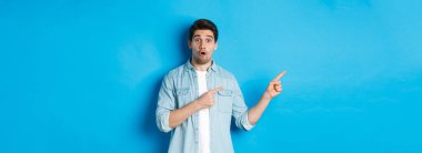Gündelik giysiler içinde şaşırmış yetişkin bir adamın portresi duyuru yapıyor, parmaklarını sola gösteriyor ve şaşkın şaşkın bakıyor, mavi arka planda duruyor..