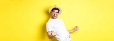 Turizm ve tatil kavramı. Tatmin olmuş genç erkek turist bir şey kazanıyor ve seviniyor, yumruk tokuşturuyor ve bağırıyor, sarı arka planda duruyor..