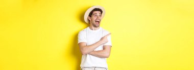 Turizm ve yaşam tarzı kavramı. Hasır şapkalı heyecanlı yakışıklı adam reklama bakıyor, sol üst köşedeki logoya bakıyor, sarı arka plan..