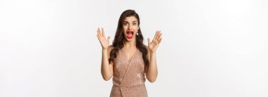 Kutlama ve parti konsepti. Zarif elbiseli güzel bir kadın çığlık atıyor ve kameraya bakıyor korkmuş, korkunç bir şeye bakıyor, beyaz arka planda duruyor..