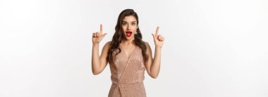 Noel, tatiller ve kutlama konsepti. Parti elbisesi içinde güzel bir genç kadın, hayret içinde, parmaklarını havaya kaldırarak reklamını yapıyor, en iyi teklifi veriyor, beyaz arka plan.