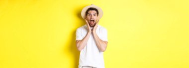 Turizm ve yaz kavramı. Hasır şapkalı şaşırmış erkek model, sarı arka planda duran, özel tekliflere şaşırmış görünüyor..