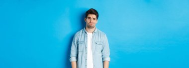 Kameraya şüpheyle bakan, somurtan, somurtan, bir şey hakkında huzursuz hisseden, mavi arka plana karşı duran bir adam..