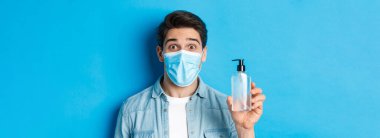 Covid-19, salgın ve karantina kavramı. Tıbbi maskeli, el dezenfektanı şişesini tutan, kaşlarını kaldıran, mavi arka planda duran şaşırmış bir adam..