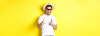 Tatilde seninle flört eden, kamerayı işaret eden ve göz kırpan güneş gözlüklü, sarı arka planda yaz şapkası takan kendinden emin ve küstah bir adam..