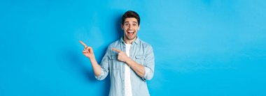 Heyecanlı, sıradan giyinmiş, reklamını yapan, fotokopi alanını işaret eden ve gülümseyen, mavi arka planda duran yakışıklı bir adam..