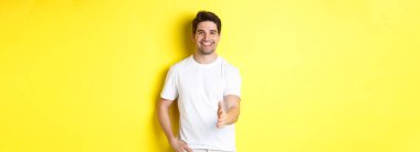 Yakışıklı ve kendine güvenen bir adam el sıkışmak için elini uzatıyor, seni selamlıyor, merhaba diyor, sarı arka planda beyaz tişörtle dikiliyor..