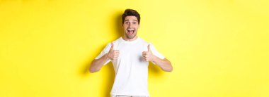 Heyecanlı yakışıklı adam başparmağını kaldırıyor, onaylıyor ve evet diyor, sarı arka planda duruyor..