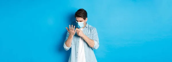 Concept of coronavirus, social distancing and pandemic. Man in medical mask looking at his palm through magnifying glass, standing over blue background.