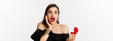 Kırmızı rujlu, makyajlı, evlilik teklifi aldıktan sonra şaşırmış görünen, nişan yüzüğünü tutan, beyaz arka planda duran çekici genç bir kadın..