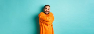 Mutlu adam kendini rahat hissediyor, sıcak bir süveter giyiyor ve kendine sarılıyor, gülümsüyor, açık mavi arka planda duruyor..