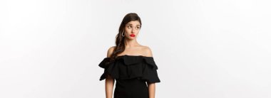 Moda ve güzellik. Kafası karışmış, siyah elbiseli, kafası karışmış ve şok olmuş bir halde beyaz arka planda duran çekici bir kadın..