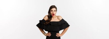 Güzellik ve moda konsepti. Siyah elbiseli zarif bir kadın kameraya bakıyor. Şaşırmış, tamamen güvensiz görünüyor. Beyaz arka planda şaşkın şaşkın duruyor..