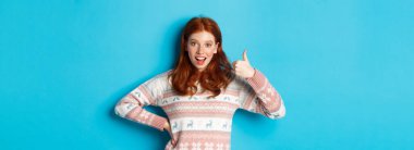 Heyecanlı kızıl kız başparmağıyla destek veriyor, gülümsüyor memnuniyet içinde, evet diyor, kabul ediyor, mavi arka planda duruyor..