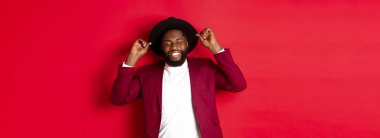 Noel alışverişi ve insanların konsepti. Mutlu siyah adam yeni yılı kutluyor, parti kıyafeti giyiyor, şapkaya dokunuyor ve gülümsüyor, kırmızı arka planda duruyor..