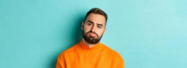 Üzgün, hayal kırıklığına uğramış ve sıkıntılı görünen, turkuaz arka planda turuncu süveter giyen üzgün genç adamın yakın çekimi..