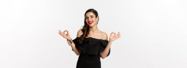 Moda ve güzellik. Siyah elbiseli çekici esmer kadın, iyi işaretler gösteriyor ve gülümsüyor, tatmin oluyor, onaylıyor ve tavsiye ediyor, beyaz arka planda duruyor..