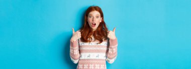 Başparmakları havada, ağzı açık, etkileyici, onaylı ve ürün gibi giyinen etkileyici kızıl saçlı kız, bir şeyleri övüp duruyor, mavi arka planda duruyor..